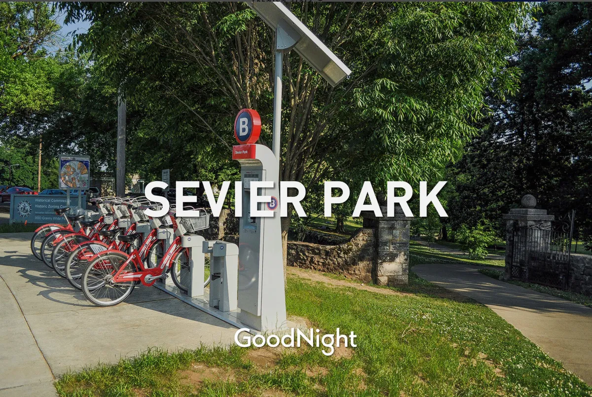 A lively scene at a local park, with a bike rental station, clear park signage, and surrounding greenery. The space is clearly designed for outdoor activities like biking, walking, and picnicking, with an energetic community vibe.