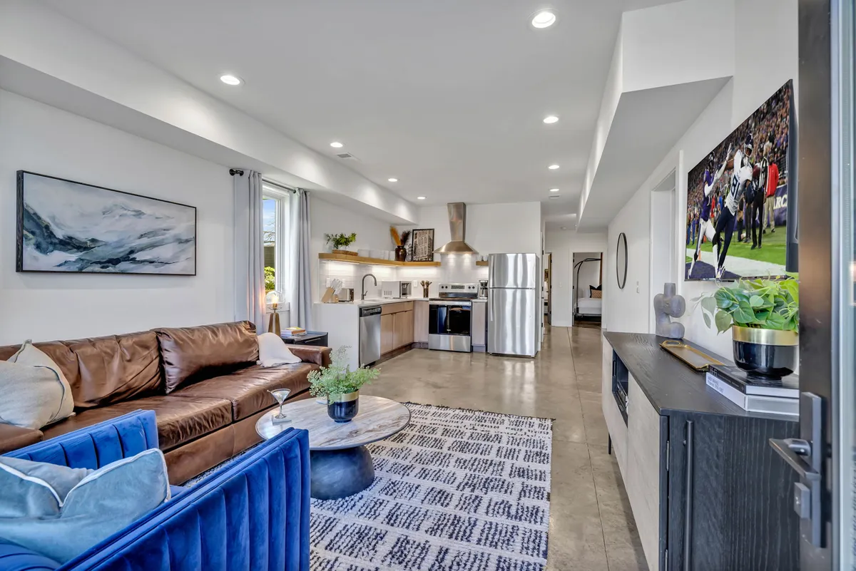 This image captures a cozy open-concept living area with a brown leather sofa, blue accent chair, round coffee table, wall-mounted TV, and a visible kitchen area in the backdrop. The room is modern yet inviting, ideal for socializing, relaxing, or enjoying entertainment.