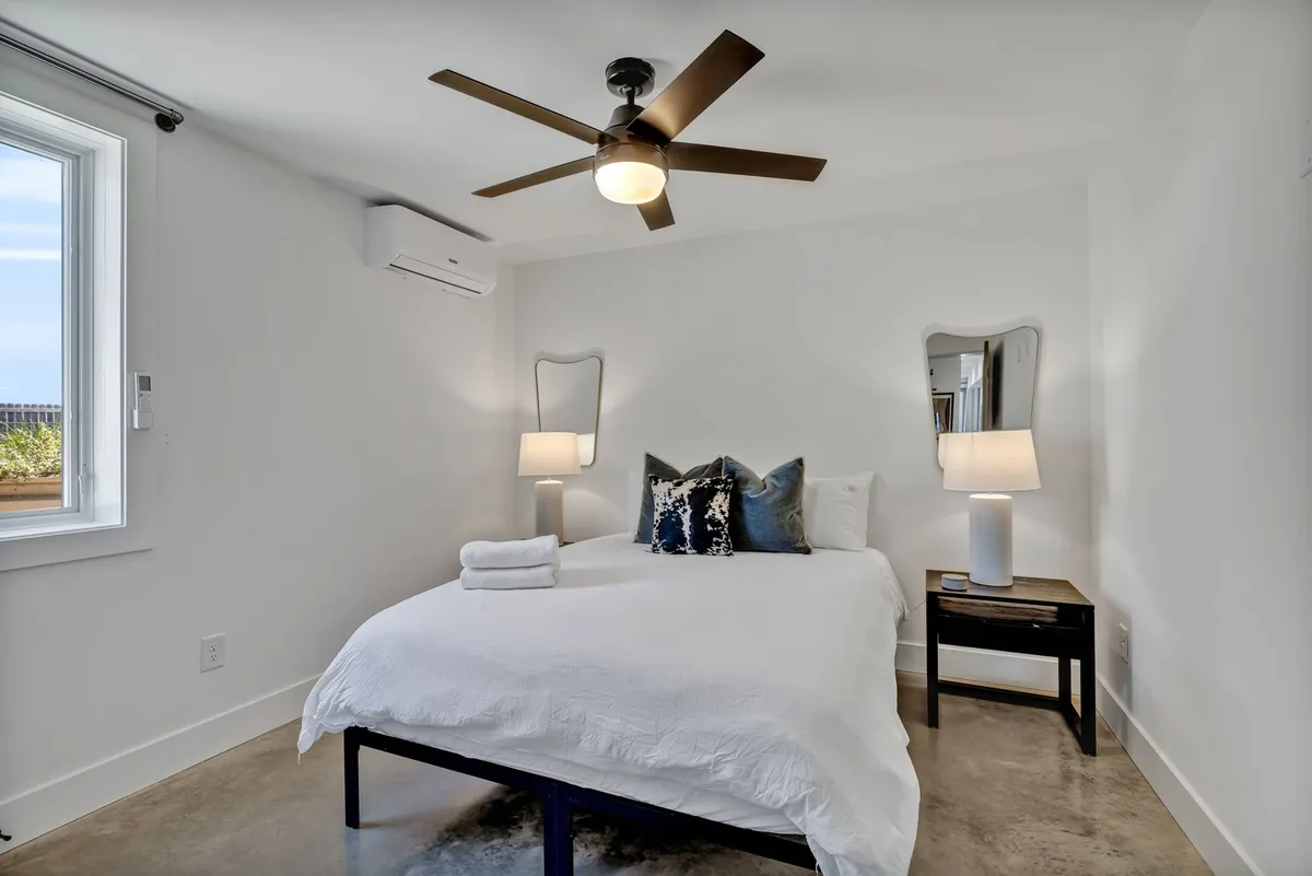 A bedroom with crisp white linens, decorative pillows, a ceiling fan, and bedside lamps. Sunlight softly streams through the window, bathing the room in a gentle glow, suggesting a quiet, restful hideaway.
