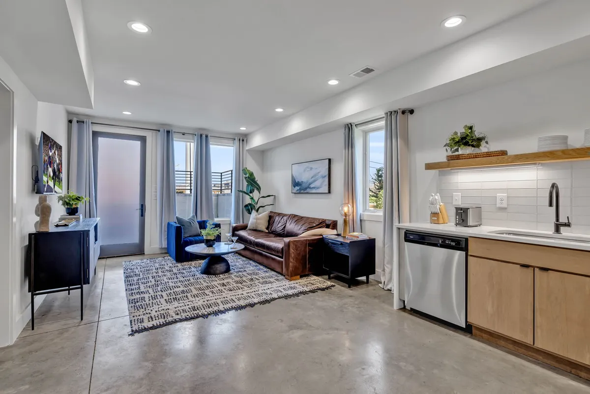 A modern, comfortable living room with a leather sofa, a blue accent chair, and a round coffee table set near a kitchen area. The space feels cozy and functional, ideal for family movie nights or casual relaxation for business guests.