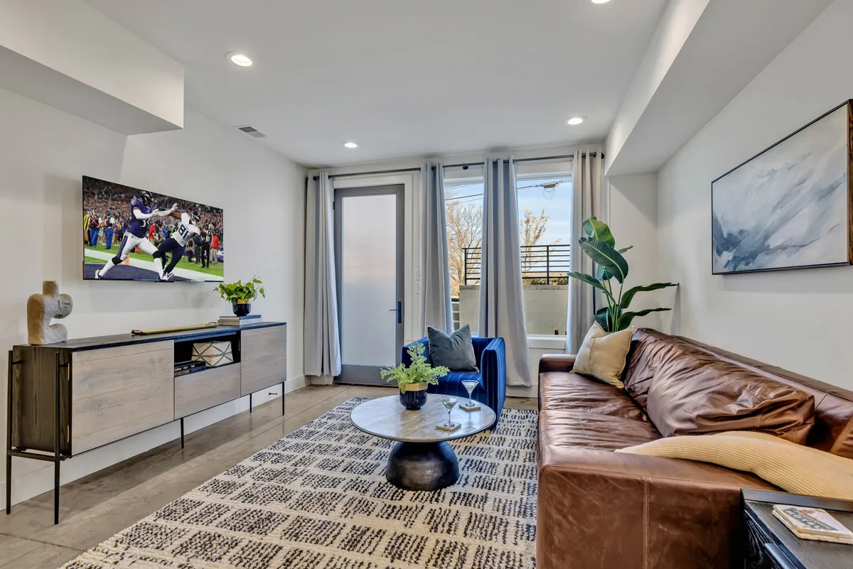The image showcases a stylish living room with designer touches, including a plush blue accent chair, a modern coffee table adorned with a plant, and a large television displaying a sports scene. The atmosphere is inviting and perfect for gathering with friends to watch the game, unwind, or enjoy intimate moments.