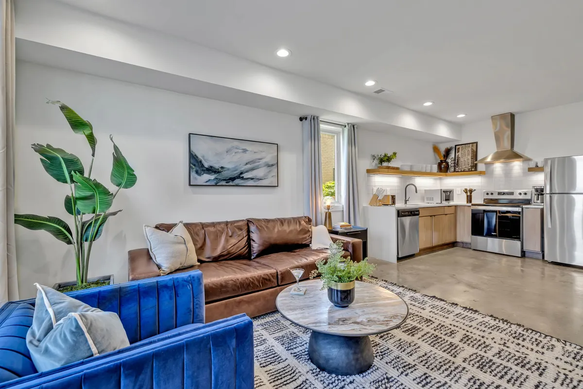 A spacious, stylish interior featuring a brown leather sofa and a blue velvet sofa, a round coffee table, modern kitchen area with contemporary appliances, abstract wall art, and a potted plant. The space is open and inviting, suitable for family gatherings, socializing, dining, and unwinding after work or travel.
